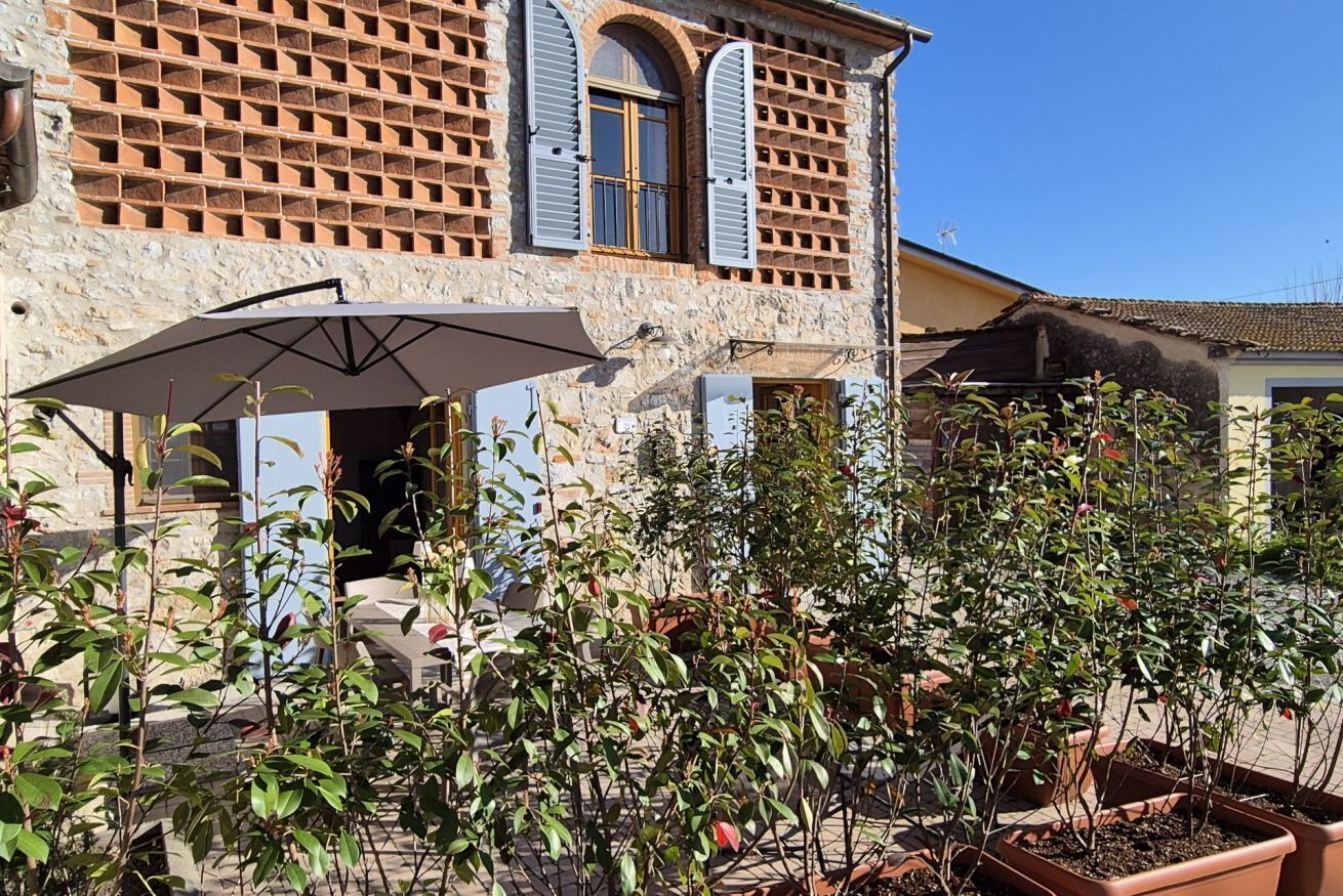 A stone building with a tiled roof, a covered outdoor seating area with a table and chairs, and potted plants.