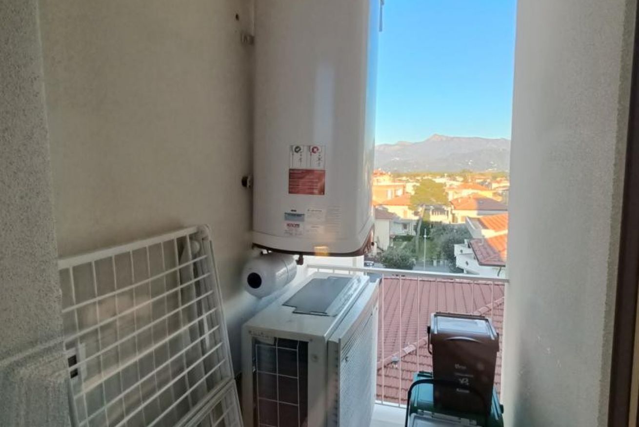 Un balcon équipé d’un chauffe-eau, d’un climatiseur et offrant une vue sur un quartier résidentiel, avec des maisons et des montagnes en arrière-plan.