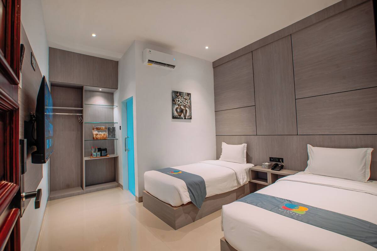 A modern hotel room with two beds, a built-in wardrobe, a small seating area, and a mounted television.