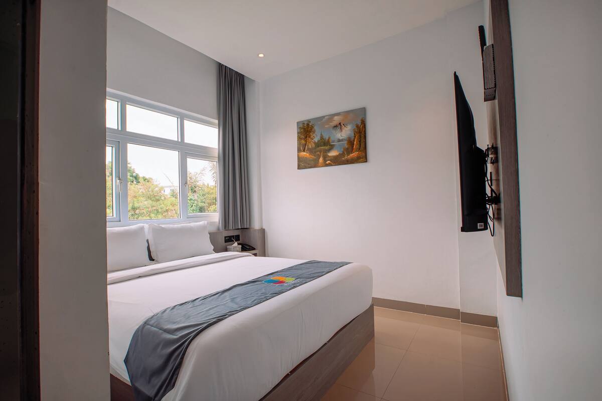 A hotel room with a large bed, a painting on the wall, a TV mounted on the wall, and a window with curtains.