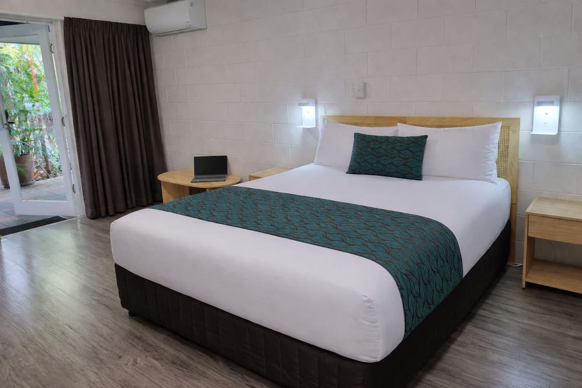 A hotel room with a bed, a laptop on a bedside table, a small table, and a door leading to a balcony.