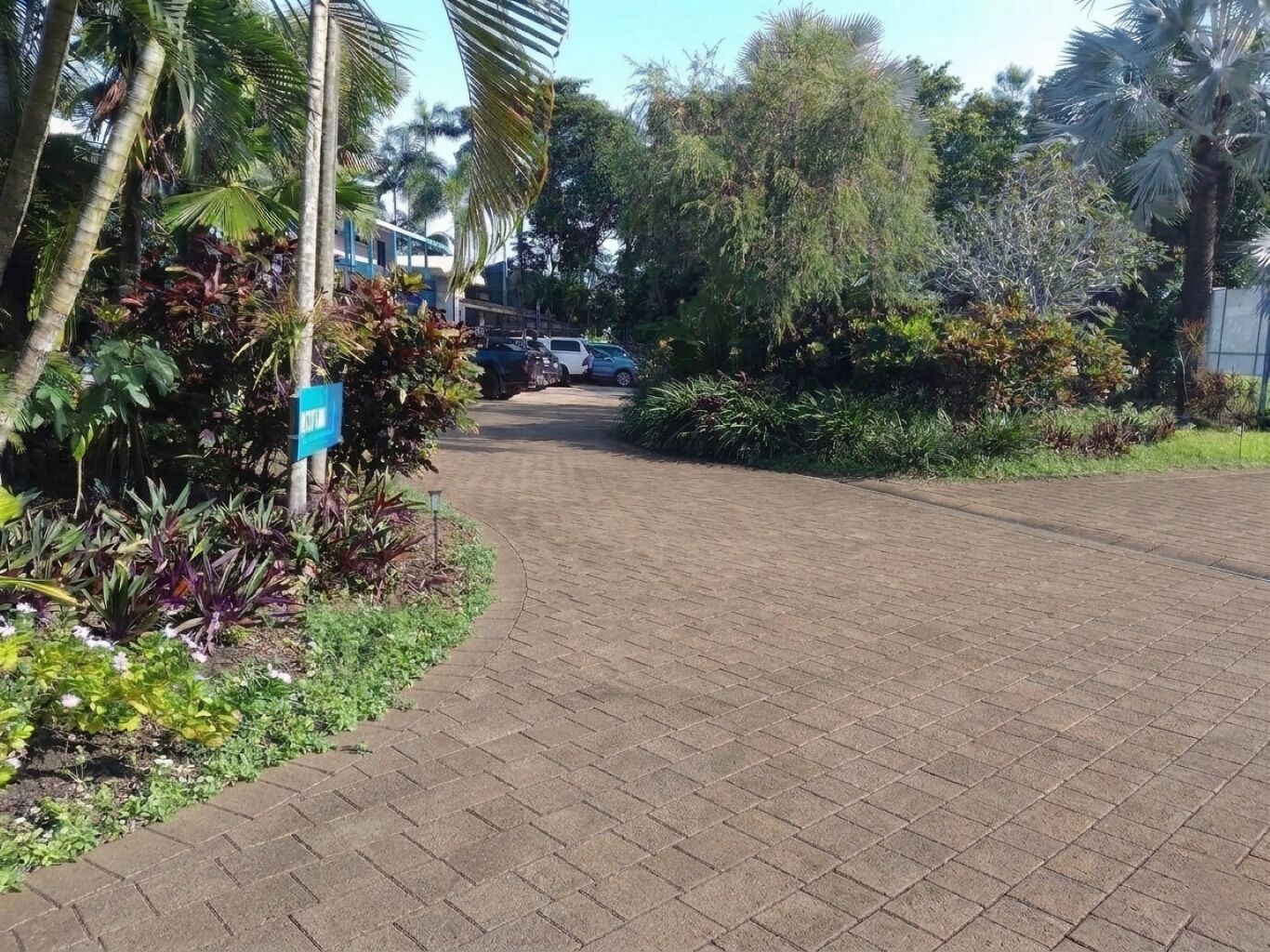 Un chemin pavé mène à un bâtiment où sont garées des voitures, le tout entouré d’une végétation luxuriante.
