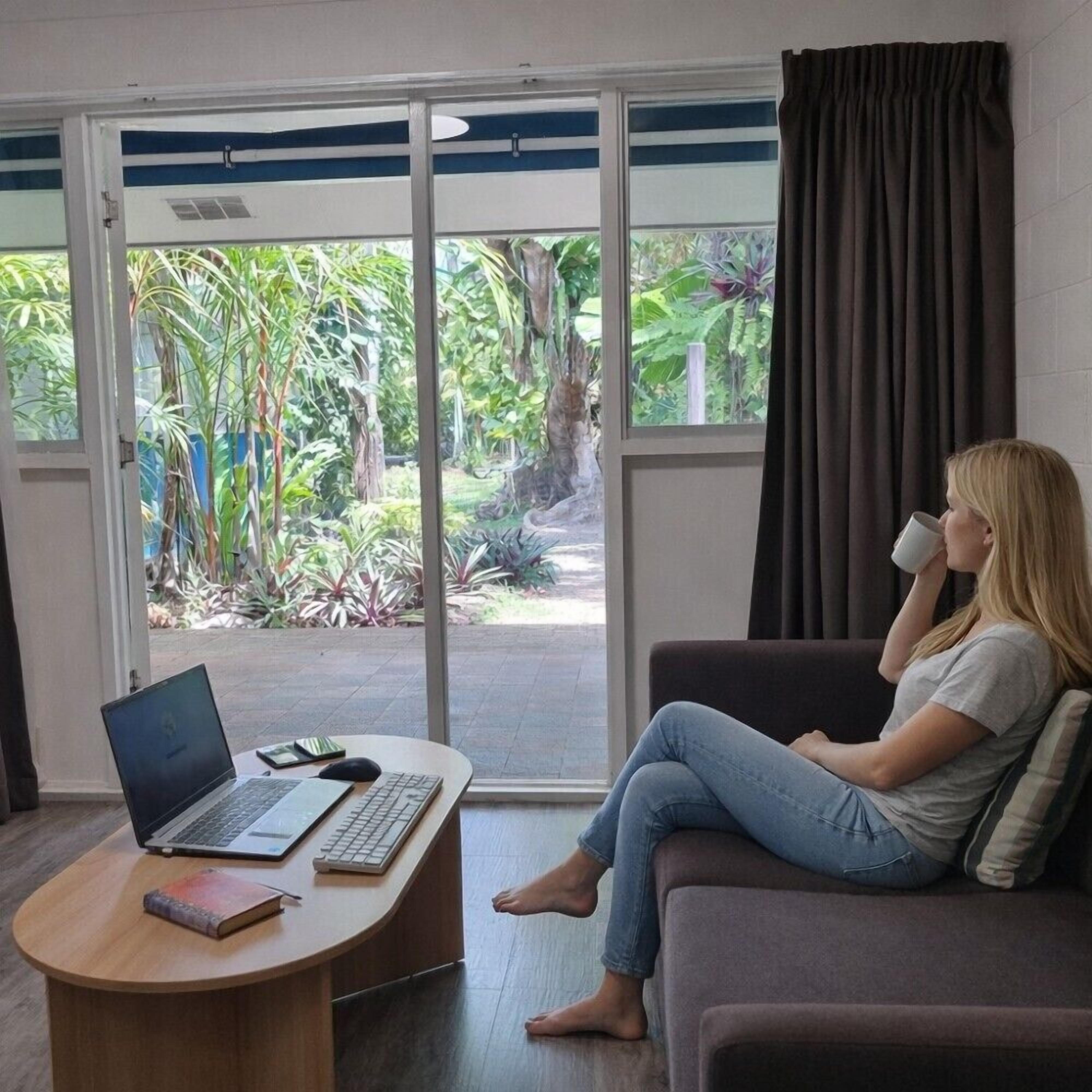 A room with a laptop on a desk, a person sitting on a couch, and a view of greenery through a glass door.