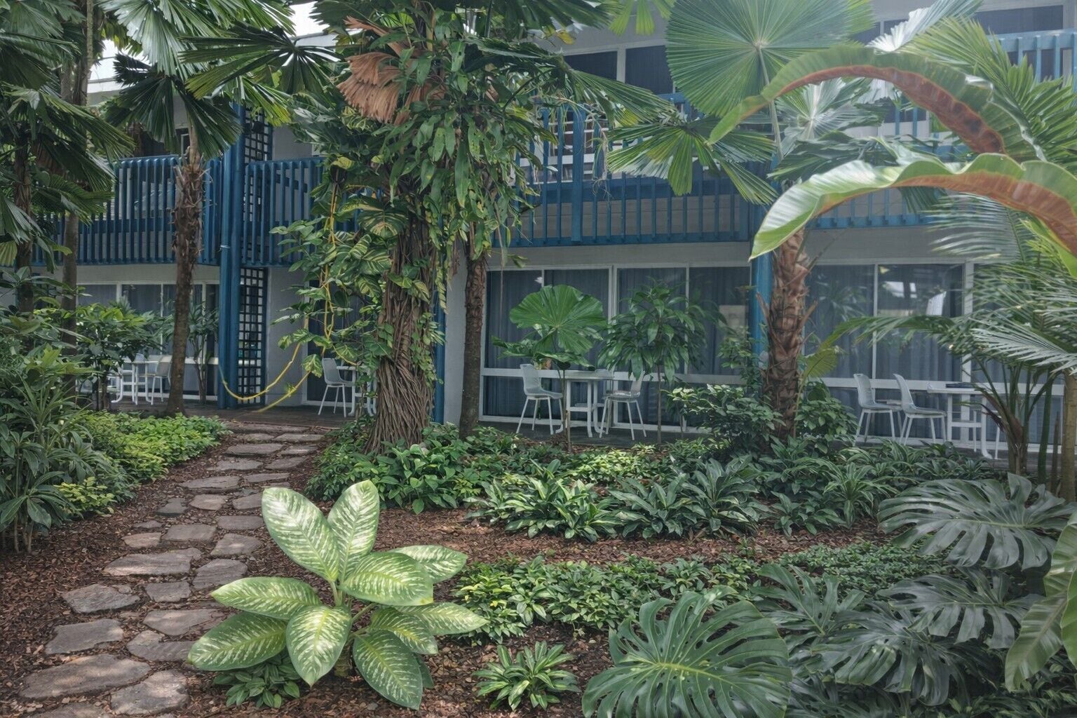 Un patio con sendero de piedra, diversas plantas tropicales, mobiliario exterior y una cerca azul.