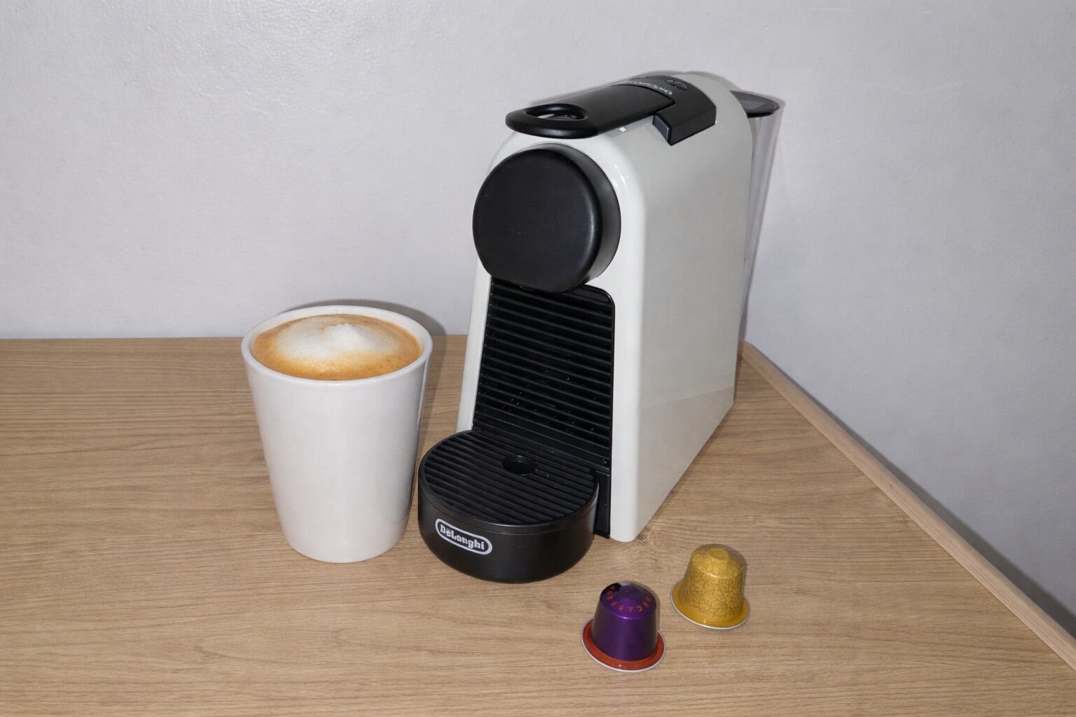 Máquina de café, uma xícara de café e duas cápsulas de café sobre uma mesa de madeira.