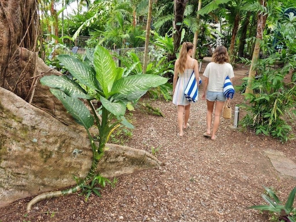 Two people walking on a gravel path in a lush garden with large green plants and a rock.