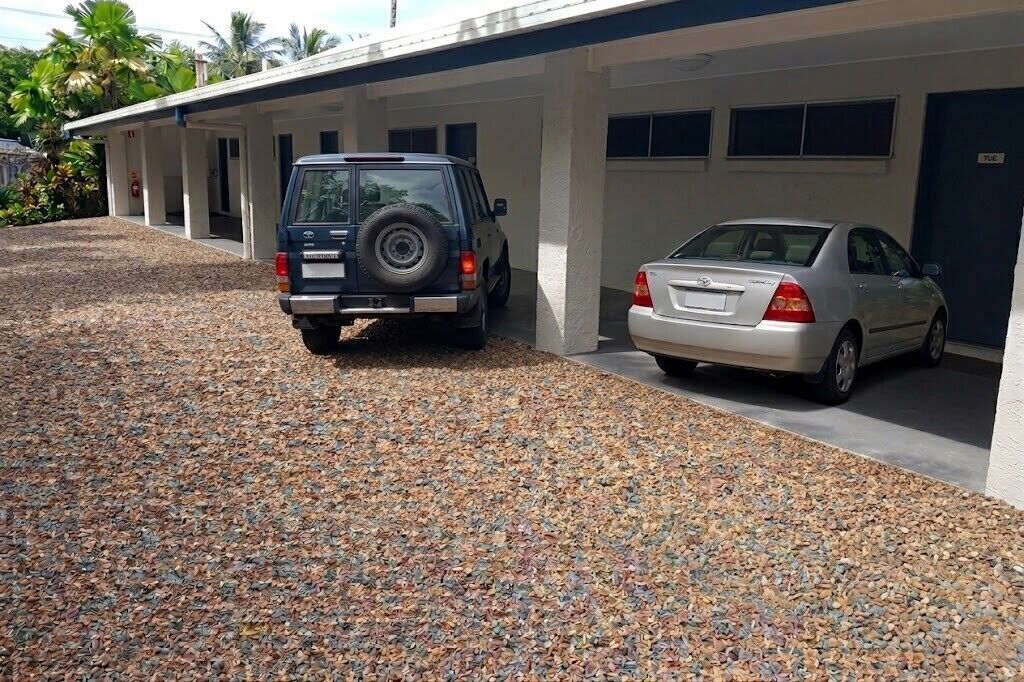 Un bâtiment doté d’un espace de stationnement couvert, d’une allée en gravier et d’une voiture blanche garée à l’intérieur.