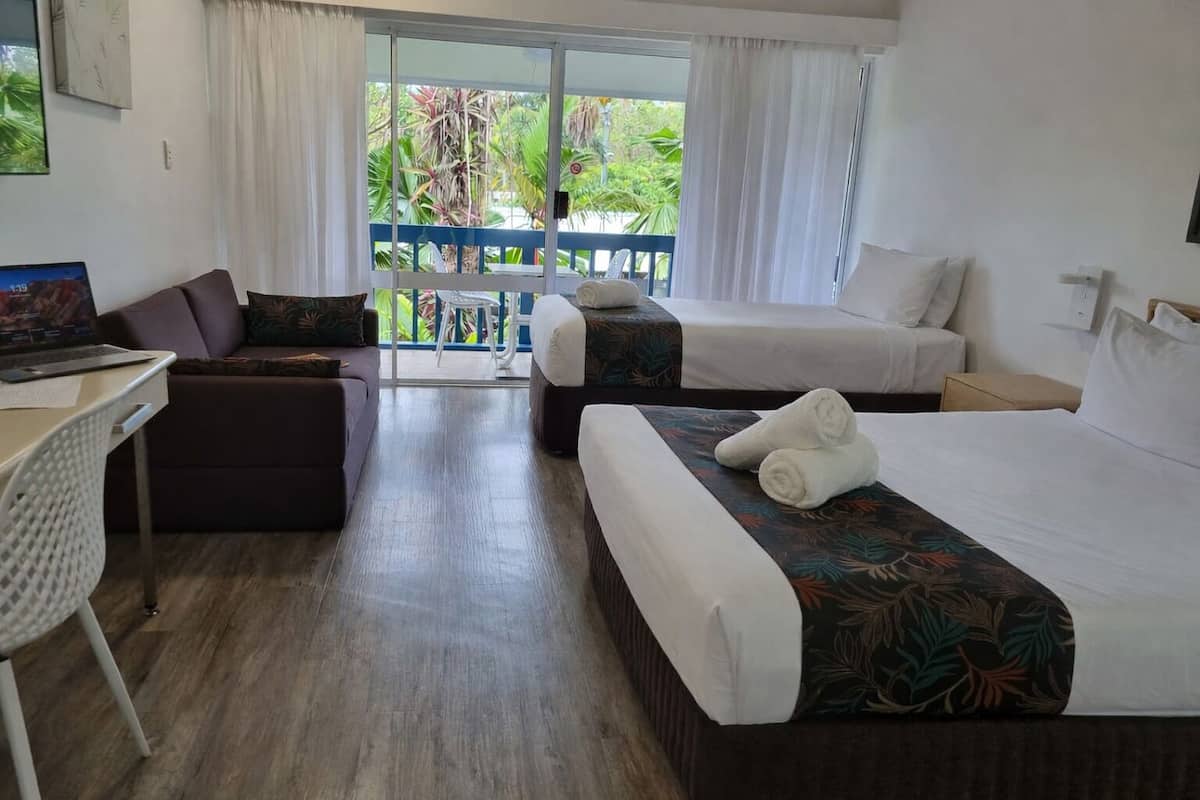 A hotel room with a desk, two beds, a sofa, and a balcony with a view of greenery.