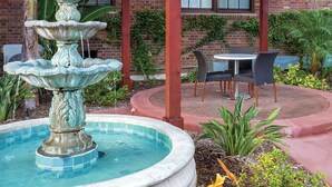 A fountain with multiple tiers in a circular pool, surrounded by a brick building with ivy and plants. There are two chairs and a small table in the background. - Comic Con 2025, San Diego, Inn at the Park 32BR (San Diego)