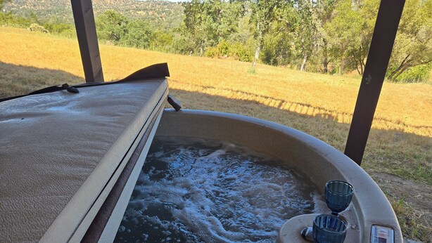 A hot tub with a wooden deck and a glass door, surrounded by a grassy field and trees. - Awesome Three-bedroom Retreat on 1.25 acres in Coarsegold with Trails and Views (Coarsegold)