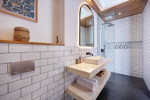 A modern bathroom with a white-tiled shower, a large mirror, and a built-in sink with a wooden shelf above. - Villa Lila (Canggu)