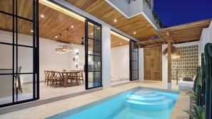 A modern indoor pool area with a wooden ceiling, glass doors, and a seating area with a table and chairs. - Villa Lila (Canggu)