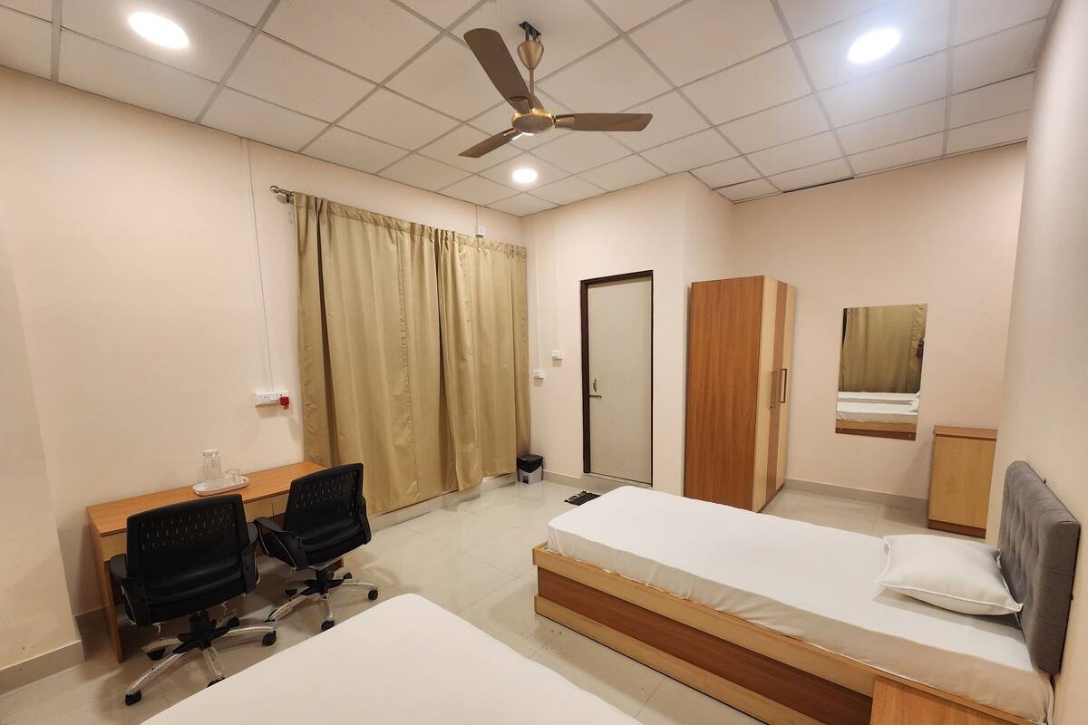 Quarto de hotel com duas camas de solteiro, uma escrivaninha com cadeira, guarda-roupa, ventilador de teto e espelho.