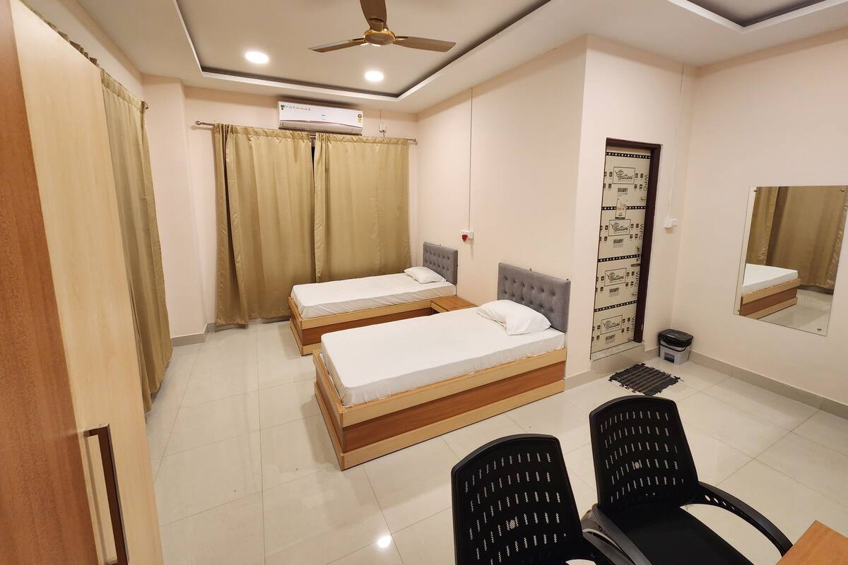 Quarto de hotel com duas camas, ventoinha no teto, ar condicionado, espelho e uma pequena lixeira.