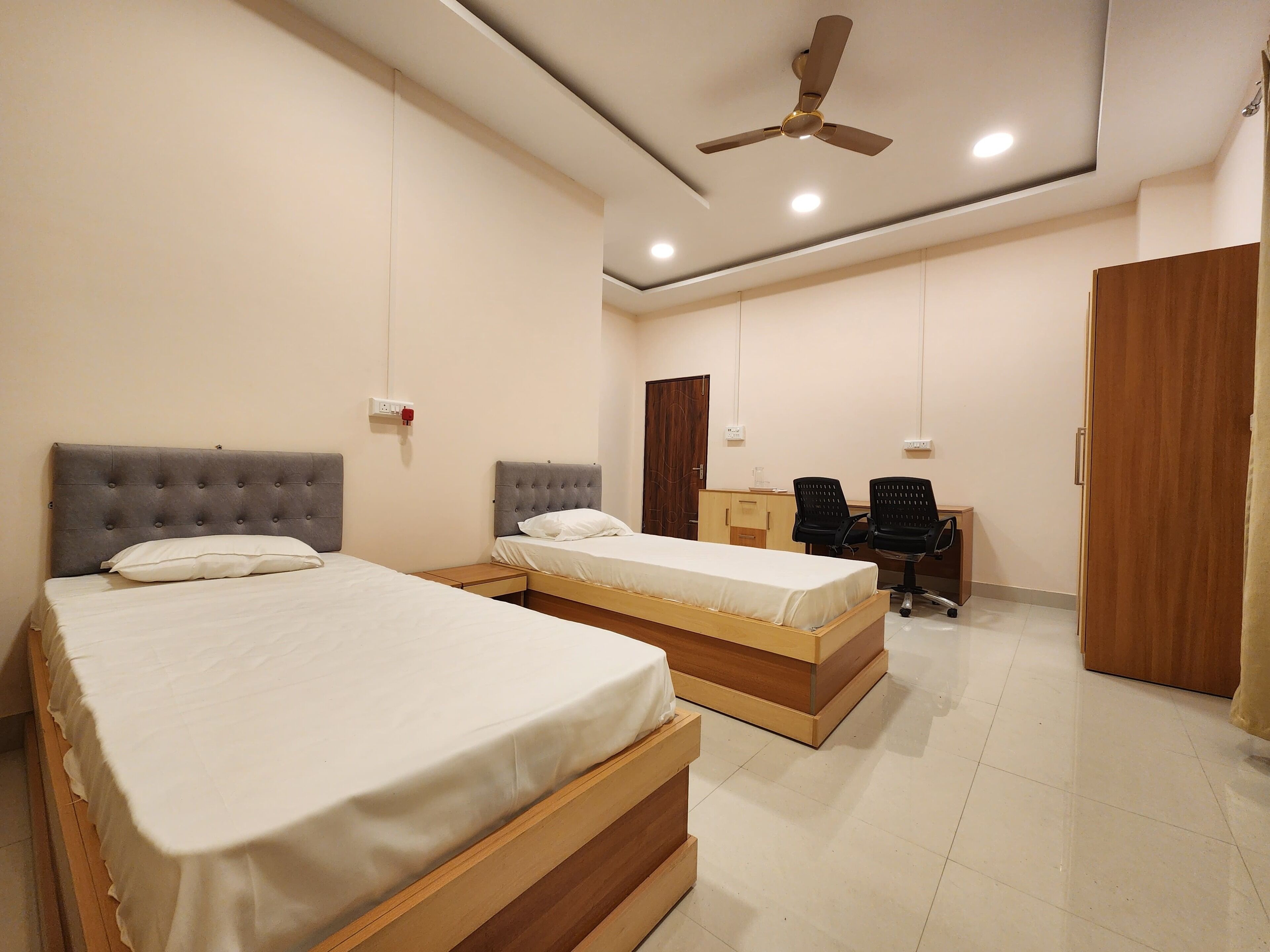 A hotel room with two beds, a desk with two chairs, a wardrobe, and a ceiling fan.