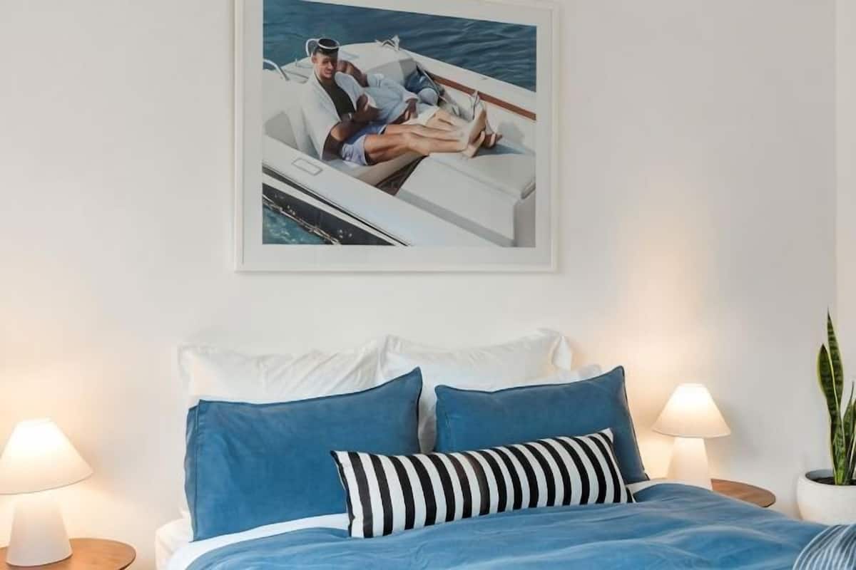 Una cama con ropa de cama azul, un cuadro enmarcado de un barco, dos lámparas de noche y una planta en maceta.