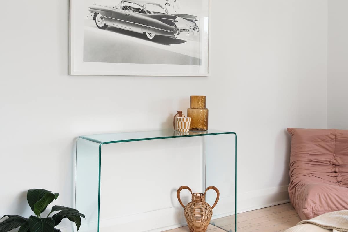 Una habitación moderna con una mesita de cristal, un grabado en blanco y negro de un coche, una planta en maceta y una cama rosa.
