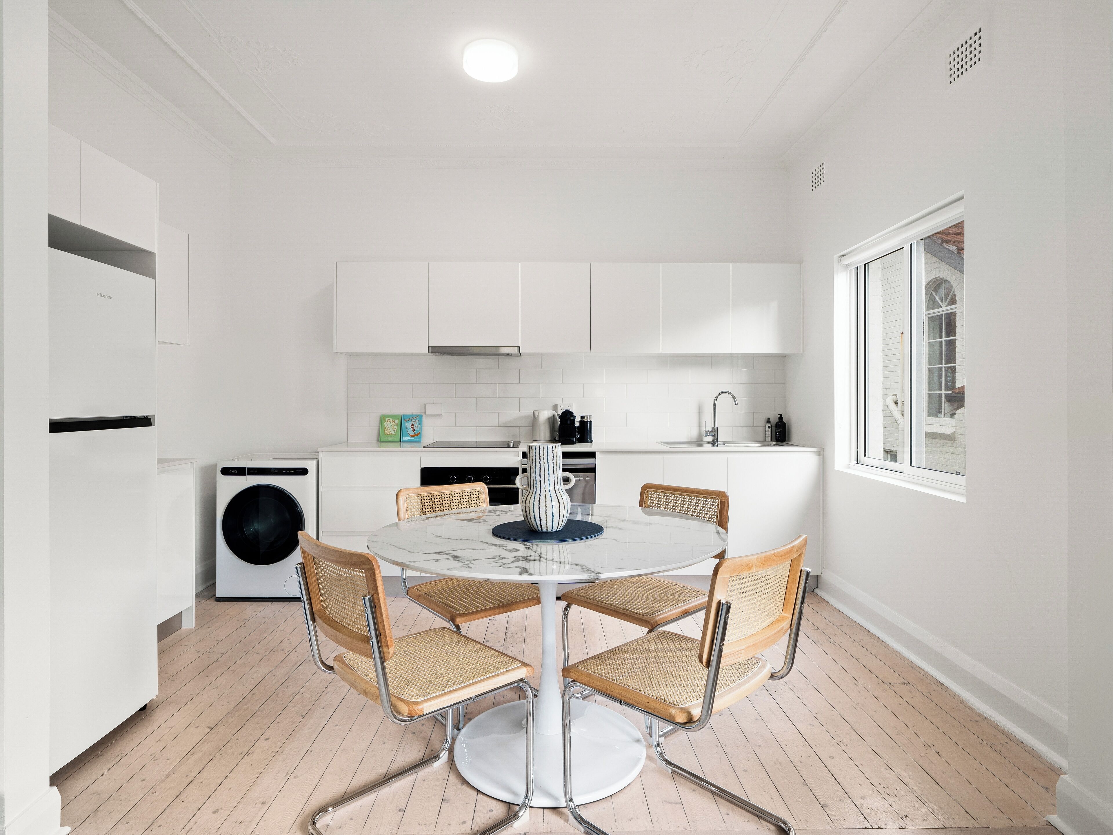 A modern kitchen with white cabinetry, a built-in washing machine, and a dining area with a round table and wicker chairs.