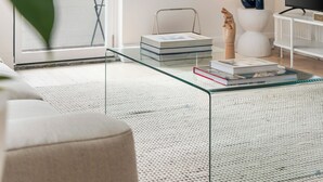 Un salon moderne avec une table basse en verre, une pile de livres, une sculpture en bois, un téléviseur et une lampe.
