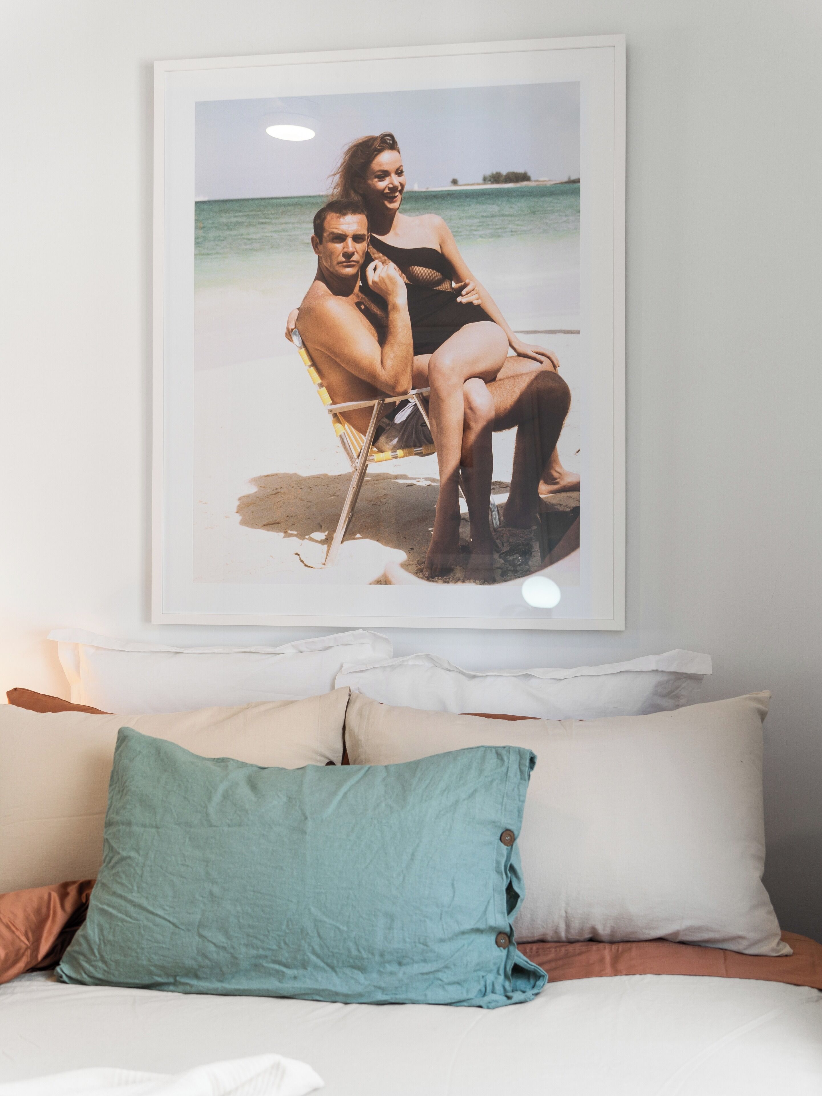 A framed photograph of a couple on a beach, a bed with white and teal pillows, and a white wall.