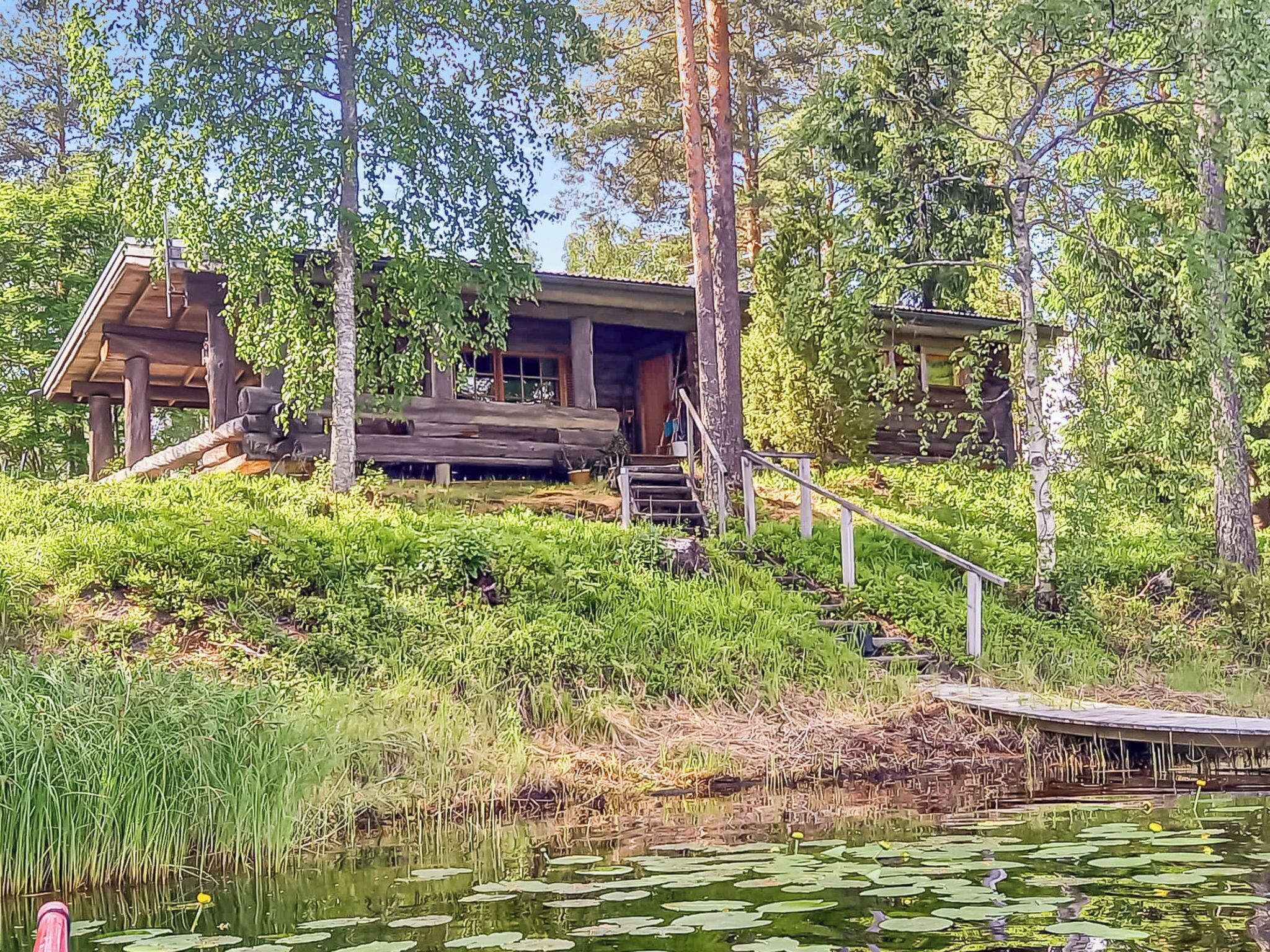 Mökki, jossa on puinen terassi, ja sen ympärillä on tiheää kasvillisuutta sekä lampi, jonka pinnalla kelluu lumpeenlehtiä.