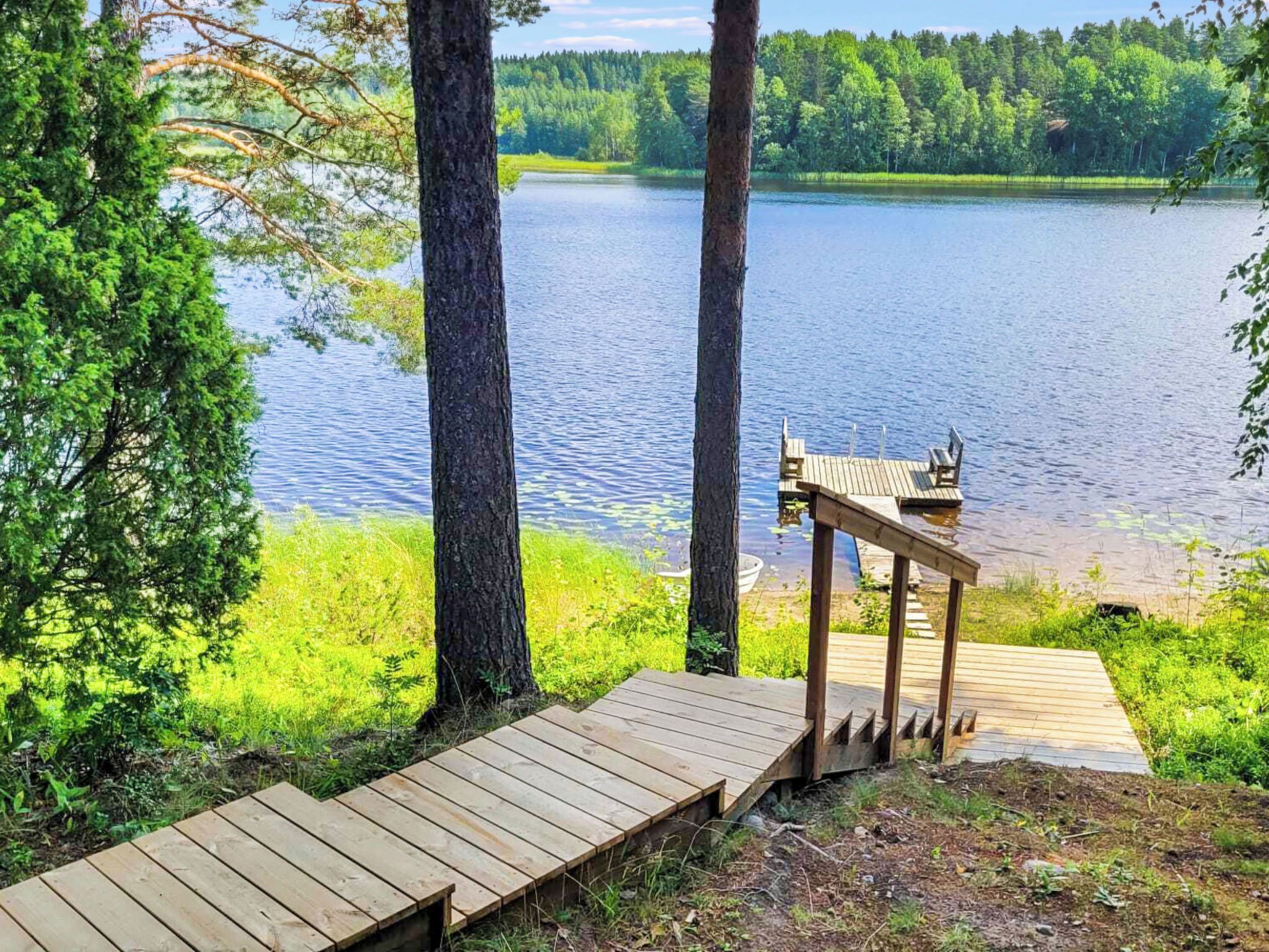 Puinen terassi, joka johtaa laiturille tyynen järven ylle. Terassin ympärillä on puita ja vehreää kasvillisuutta.
