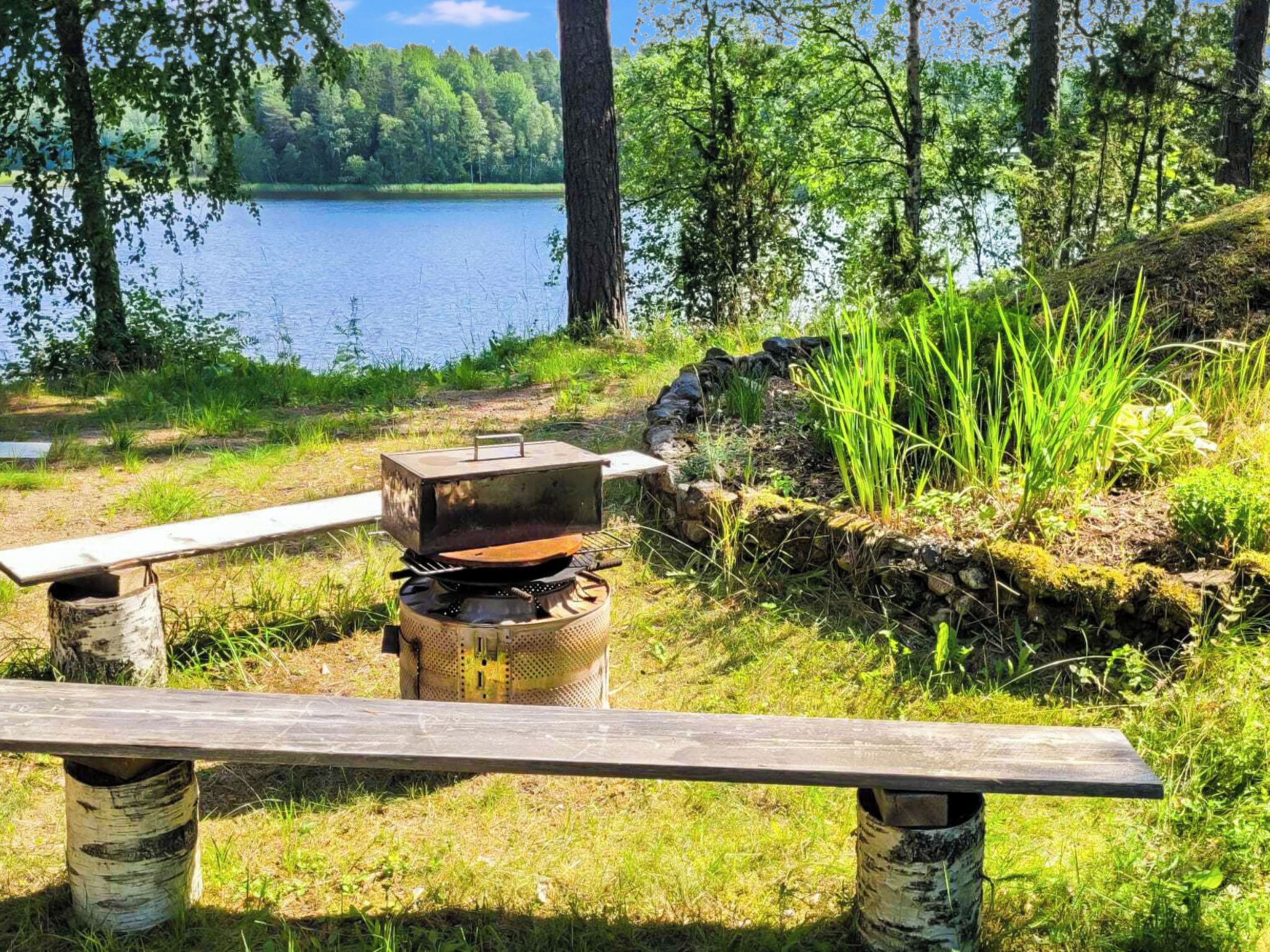 A camping setup with a portable grill and a bench, surrounded by a natural landscape with a lake and trees.