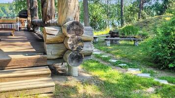 Una terraza de madera con un área de estar al aire libre de estilo rústico, una pila de leña y un sendero de piedra que lleva a una mesa de picnic.