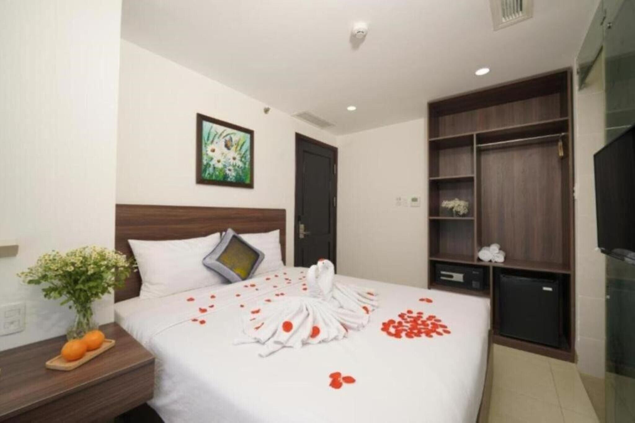 A hotel room with a neatly made bed, a bedside table with a vase of flowers, a small refrigerator, and a wardrobe.