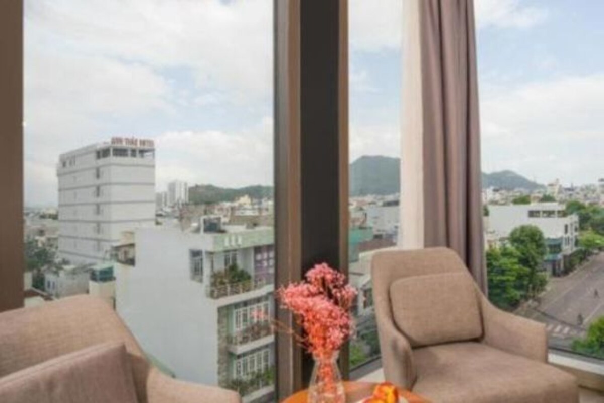Hotelkamer met een groot raam met uitzicht op de stad, een vaas met bloemen op de tafel en twee stoelen.