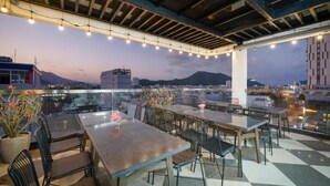 An outdoor dining area with tables and chairs, string lights, and a view of the cityscape and mountains in the background.