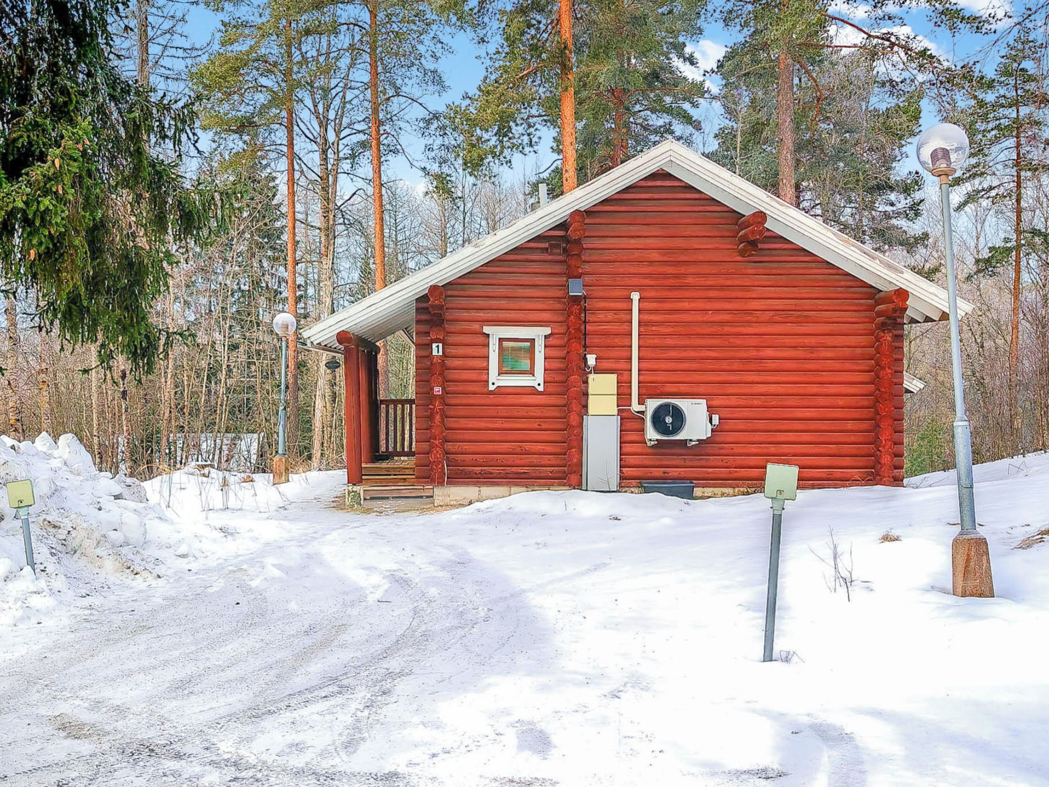 En röd timmerstuga med en enda dörr och ett litet fönster, omgiven av snö och träd.