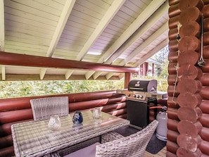 A covered outdoor seating area with a table and chairs, a grill, and a log wall.