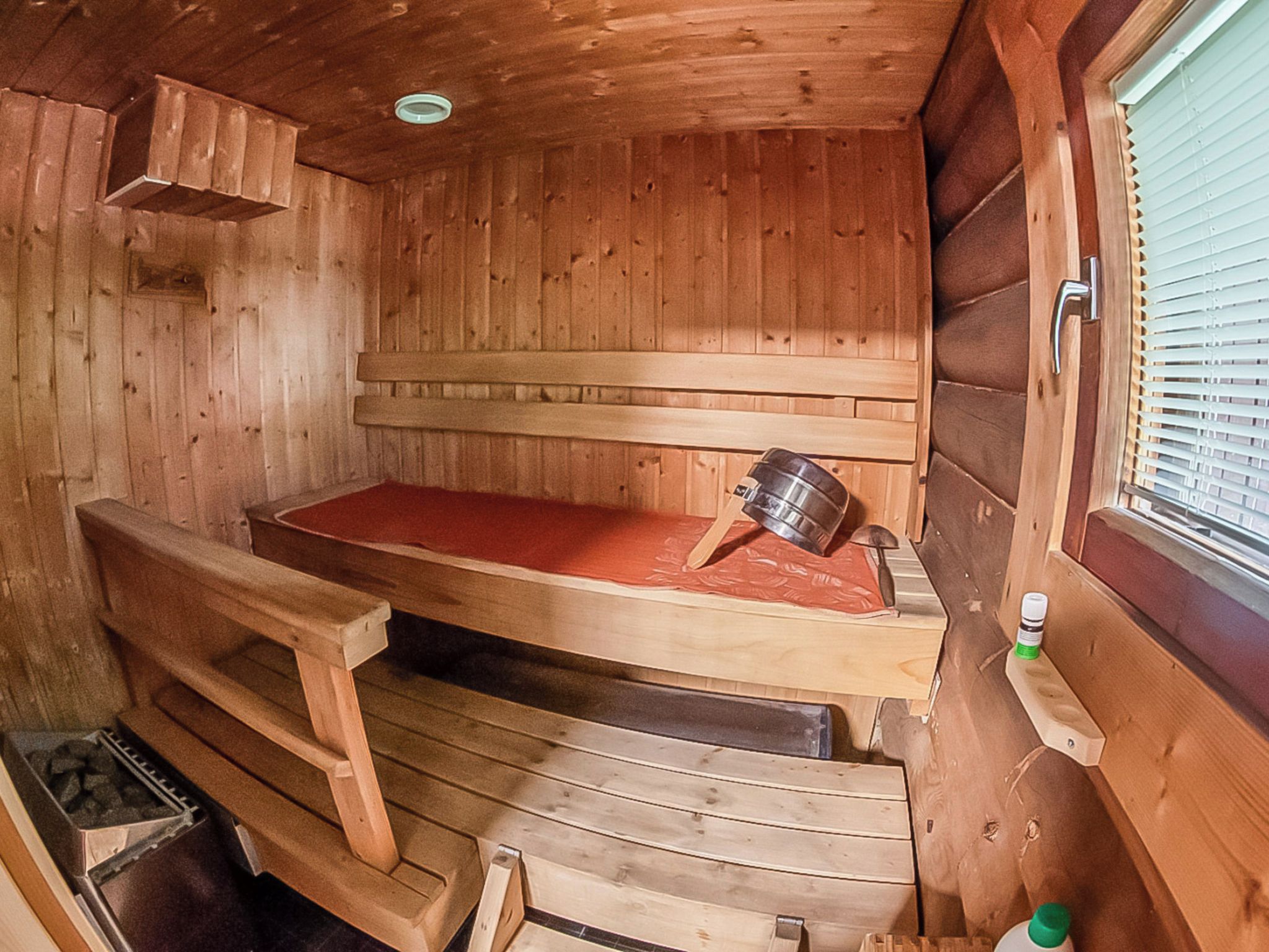 A wooden sauna with benches and a bench seat, a window with blinds, and a small shelf with a bottle.