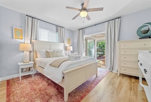 Quarto com cama, mesas de cabeceira, cômoda, ventoinha no teto e vista através de uma porta de vidro.