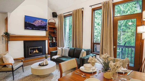 A cozy living room with a fireplace, a sofa, a dining table set, and large windows with curtains.