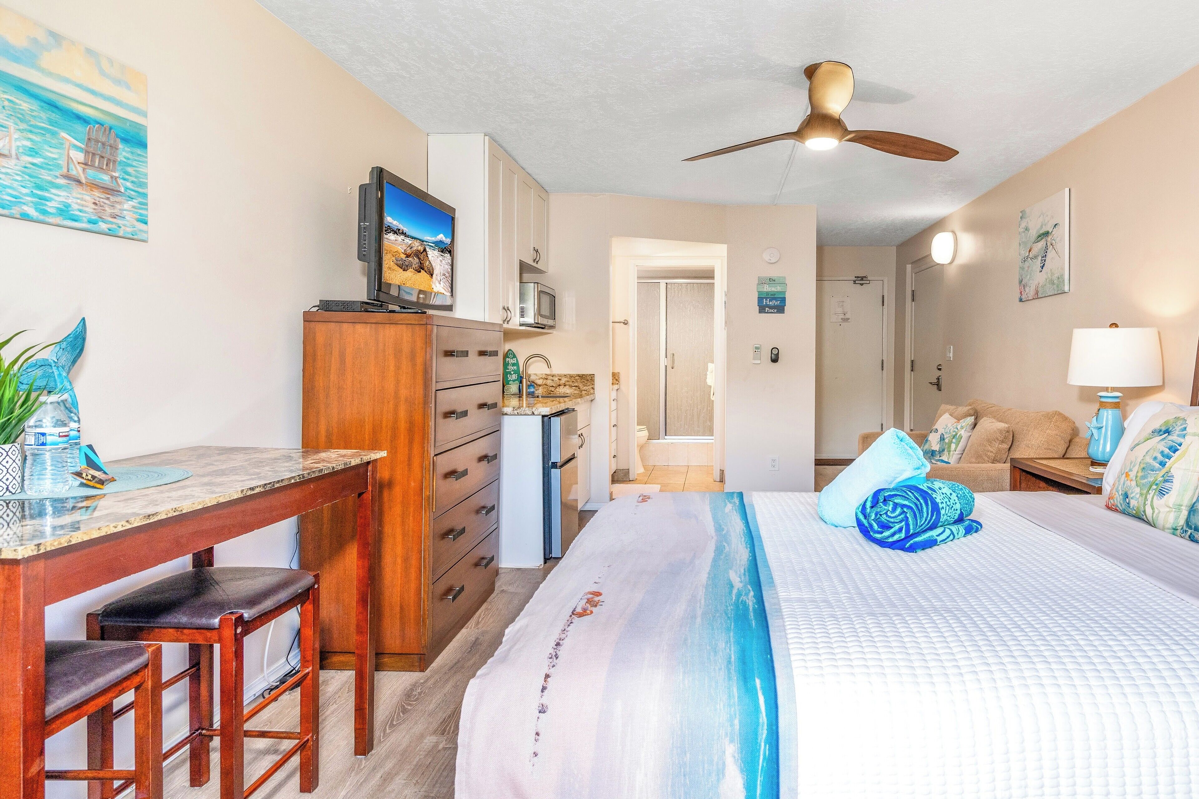 A bedroom with a bed, a desk, a chair, a ceiling fan, a TV, and a microwave.