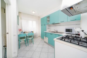A kitchen with teal cabinets, a gas stove, a microwave, and a dining area with chairs. - GuestHost - Raggio dello Scirocco Apartment - Lignano Pineta (Lignano Sabbiadoro)