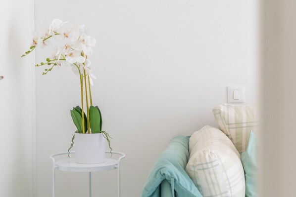 Uma orquídea branca num vaso branco sobre uma mesa branca, um sofá azul claro com almofadas xadrez e uma parede branca com um interruptor de luz.