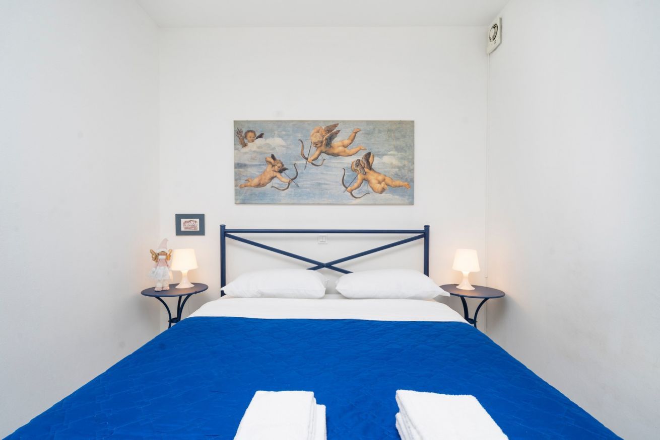 A bedroom with a bed, two bedside tables, a painting of cherubs, and a blue bedspread.
