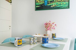 A dining table with a white tablecloth, two blue chairs, a vase with pink flowers, and a framed picture on the wall. - GuestHost - Raggio dello Scirocco Apartment - Lignano Pineta (Lignano Sabbiadoro)
