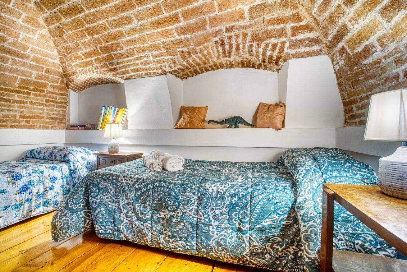 Ein Zimmer mit Ziegeldecke, einem Bett mit gemusterten Textilien, einem Nachttisch mit Lampe und einem Regal mit Büchern.
