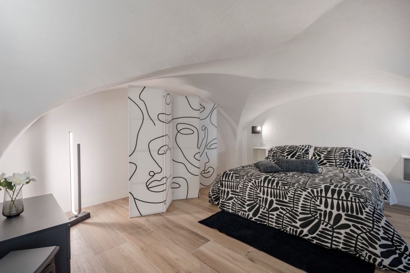 Un dormitorio con techo curvo, cama con ropa de cama estampada, alfombra negra y pared con arte lineal abstracto.