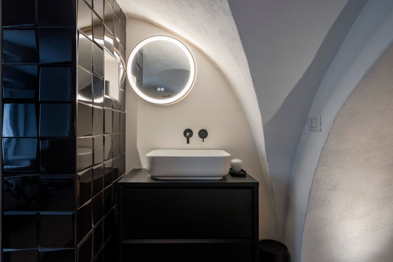 A bathroom with a white sink, a round mirror, and black tiled walls.