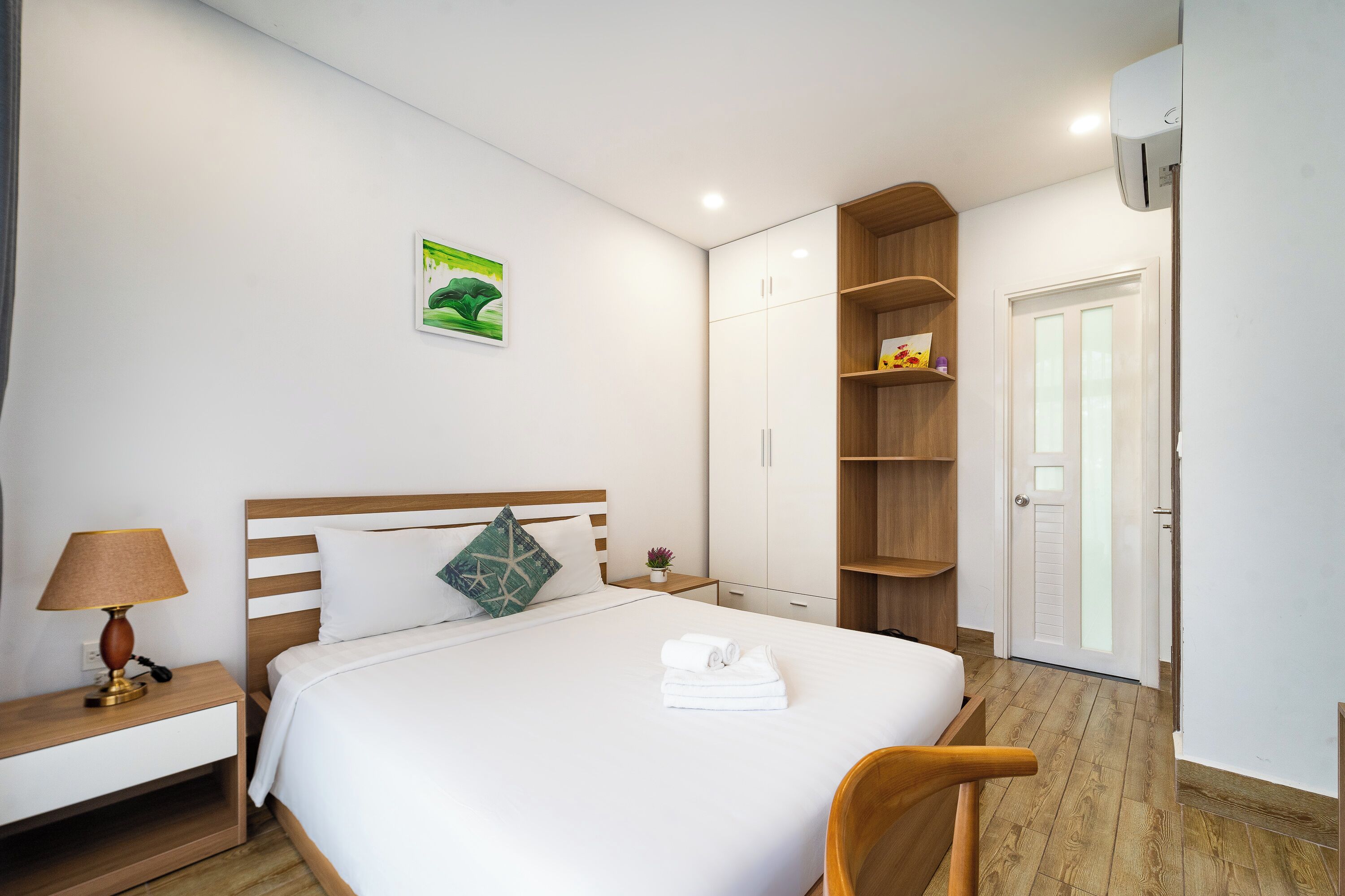A hotel room with a bed, bedside table, lamp, and a wall-mounted air conditioner.