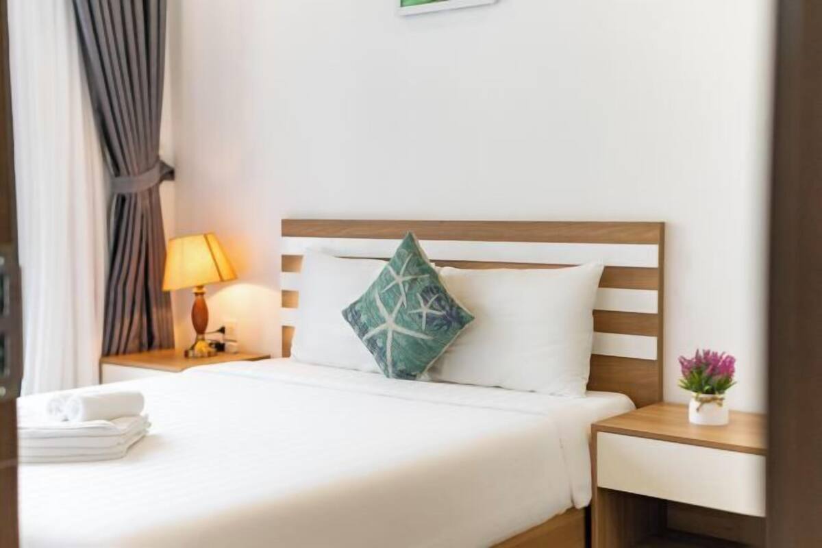 Een hotelkamer met een bed, een nachtkastje met een lamp, een kleine tafel met een bloemenvaas en een raam met gordijnen.