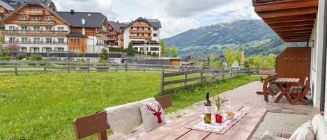 A wooden outdoor seating area with a table set for two, a bench, and a view of a mountain range.