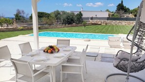 Un espace extérieur aménagé pour les repas, avec une piscine, des meubles blancs et un fauteuil suspendu.
