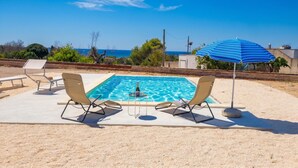 Une piscine bordée de deux chaises longues et agrémentée d’un parasol bleu, le tout entouré d’une zone sablonne offrant une vue sur la mer.