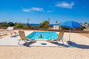 A pool with two lounge chairs and a blue umbrella, surrounded by a sandy area with a clear view of the sea.